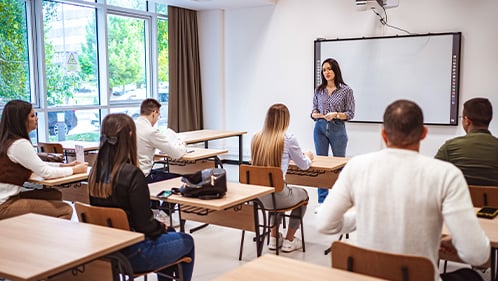 teacher stands at the front of a classroom and explains a conept to her students