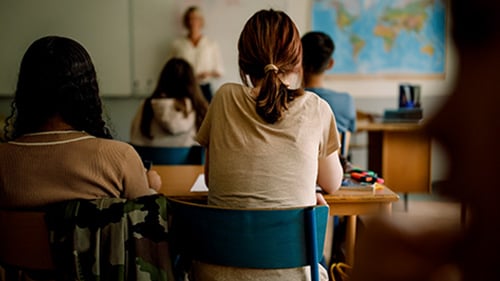 High school social studies classroom. Students face forward looking at white board and teacher.