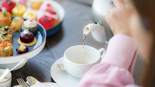 Woman pours tea into a white cup. Pastries set on an elegant plate nearby.