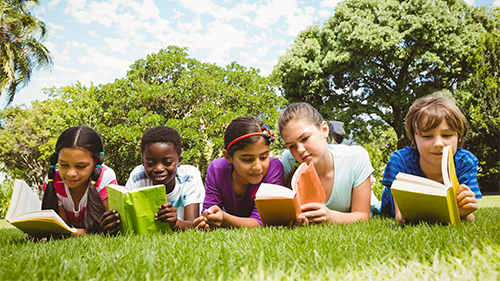 Middle school aged kids reading novels in the grass outside on a sunny day