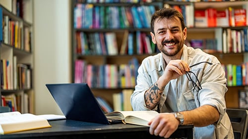 Male teacher sits at table with laptop and book open and smiles at the camera