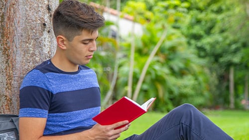 Teen boy reads book outside against a tree and appears thoughtful
