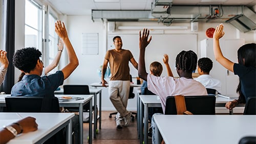 Male teacher stands at the front of a classroom of middle schoolers raising their hands