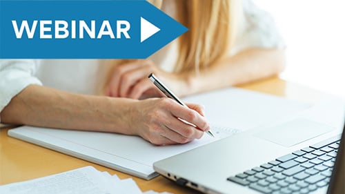 woman with blonde hair taking notes with pen and paper while looking at laptop screen