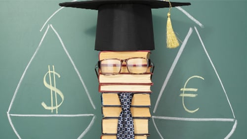 stack of books in front of chalk board. A pair of glasses, a neck tie, and graduation cap sit atop the books. 