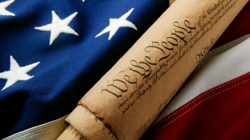 A rolled up copy of the United States Constitution sitting on top of the American flag. 