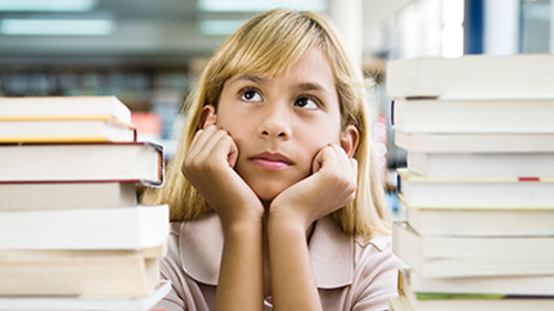 Middle school aged girl with blonde hair sitting between two large stacks of books and appearing thoughtful.