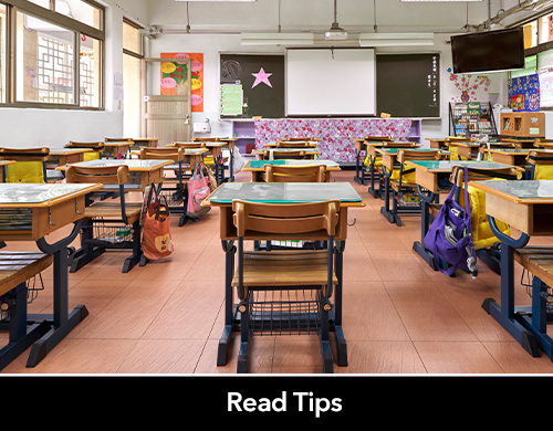 Image of Classroom with large windows and rows of desks with colorful backpacks and papers on the walls. 